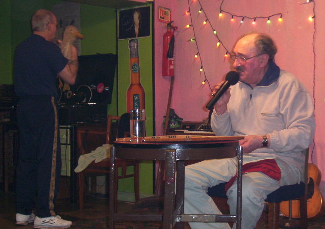 Gordon Cragg seated, placing numbers on a bingo board and talking into a chordless mike while Oz holds a ventriliquists duck.